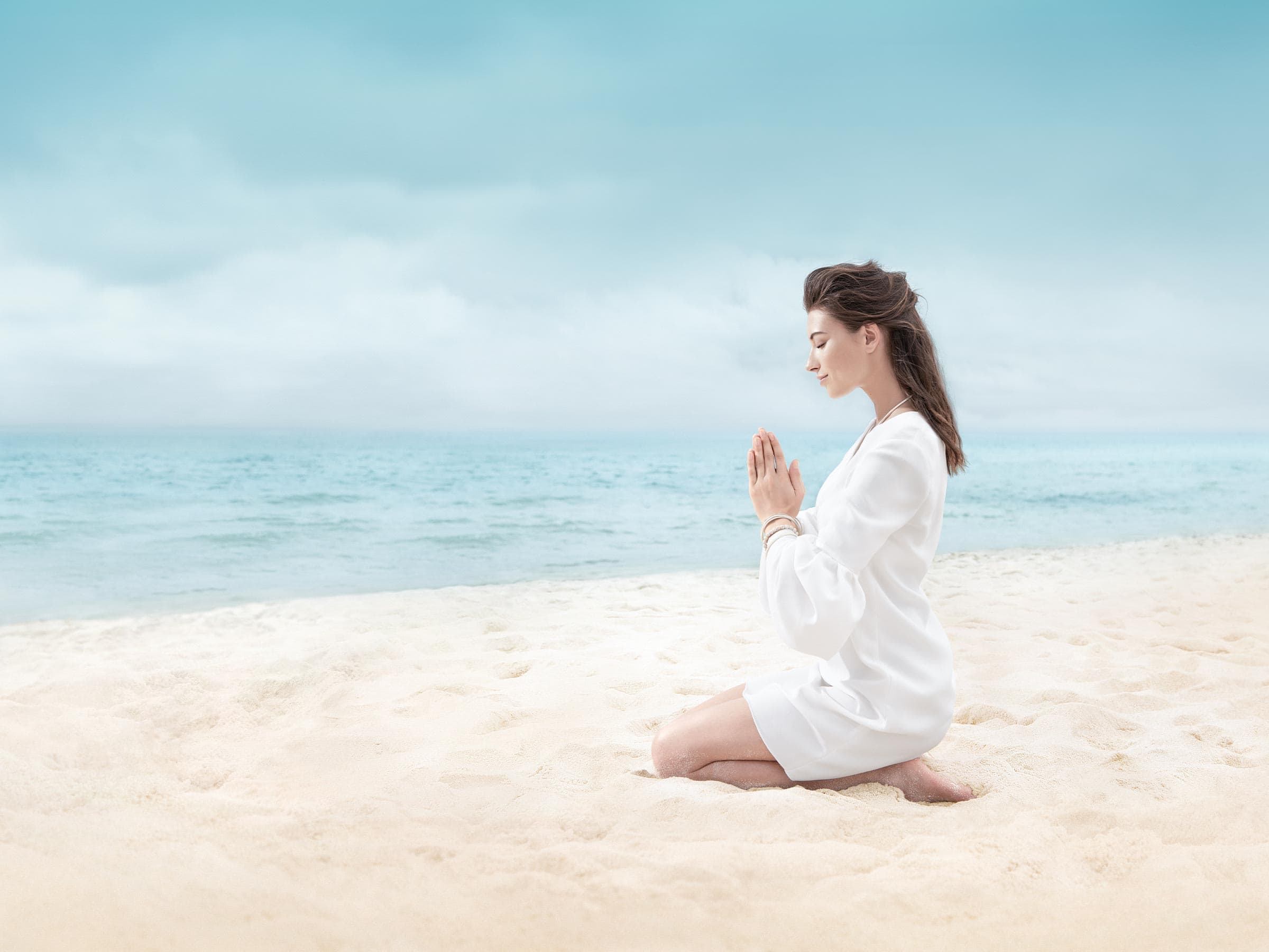 Eine Frau in einem weissen Kleid sitzt mit in Meditationspose an einem Strand
